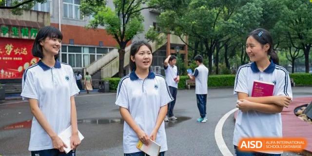 A female student from Changsha Lushan Zhongjia School wearing a white school uniform, smiling and holding papers while standing outdoors on a paved area with other students in the distance.