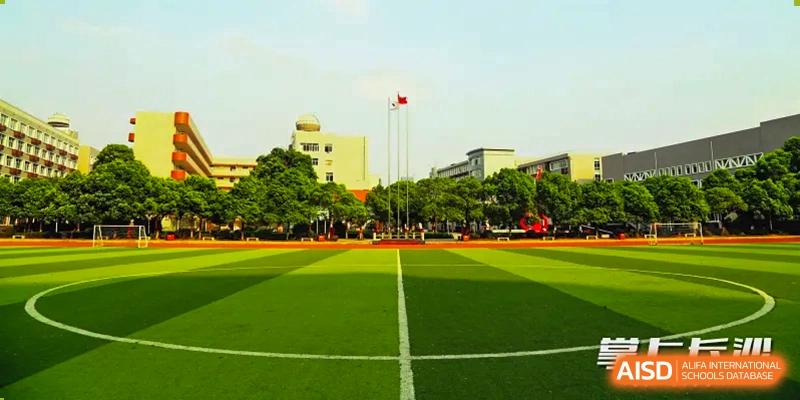 A wide-angle view of the large, well-maintained athletic field and track at Changsha Lushan Zhongjia School on a sunny day, with the main campus buildings visible in the background.