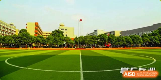 A wide-angle view of the large, well-maintained athletic field and track at Changsha Lushan Zhongjia School on a sunny day, with the main campus buildings visible in the background.
