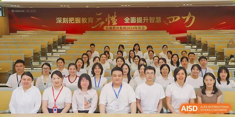 A large group of teachers and staff from Changsha Lushan Zhongjia School seated for a formal group photo in a tiered auditorium beneath a red banner with Chinese text.