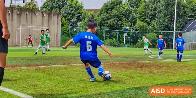A student from Changsha Lushan Zhongjia School wearing a blue jersey with the number 6, kicking a soccer ball on an outdoor field.