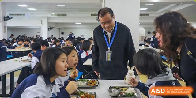 A group of students from Changsha Lushan Zhongjia School seated with an adult, all eating their meals in the school cafeteria.