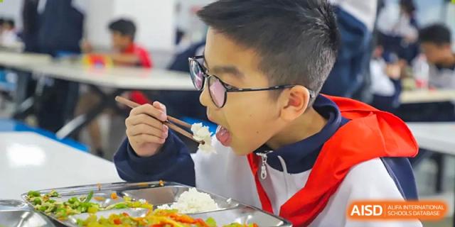 A male student from Changsha Lushan Zhongjia School wearing glasses and a red neck scarf, intently eating his meal with chopsticks.