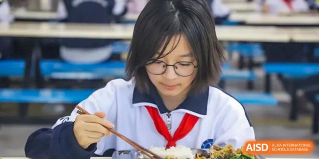 A female student from Changsha Lushan Zhongjia School wearing glasses and a school uniform, eating a meal of rice and other dishes with chopsticks.