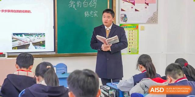 A male teacher stands in front of a classroom holding a book, at Changshu Lunhua Foreign Language School.