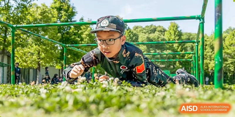 A young boy in military fatigues and glasses crawls through the grass, with Changshu Lunhua Foreign Language School.