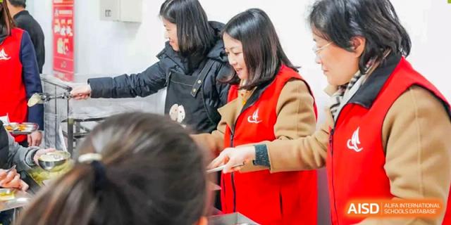 A female teacher in a red vest serves food to a student in a cafeteria line, at Changshu Lunhua Foreign Language School.