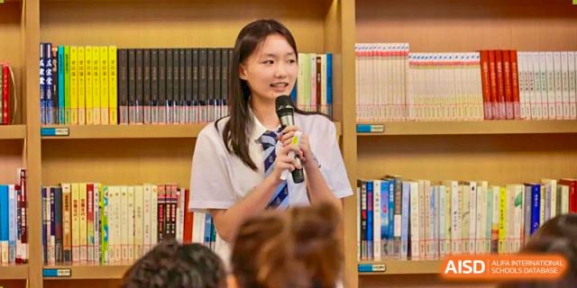 A female student from Chiway Bilingual School Xiamen speaking into a microphone while standing in front of bookshelves, addressing a group.