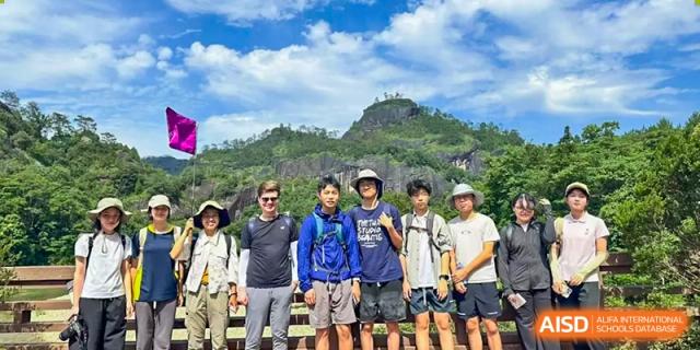 A small group of students and an adult from Chiway Bilingual School Xiamen standing in front of a large, lush green mountain during a hiking trip.