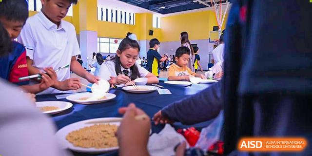 Students from Chiway Bilingual School Xiamen sitting at tables and enjoying a meal in a cafeteria or dining hall.