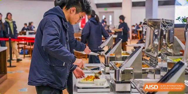 A student from Dipont Huayao Collegiate School Kunshan is serving food onto a plate from a buffet in a dining hall.