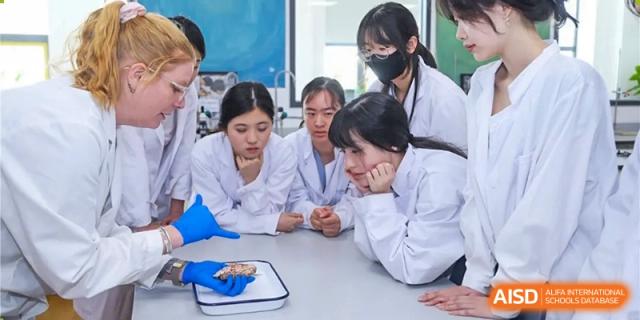 Students from Dulwich College Beijing, wearing lab coats, are gathered around a table looking at a brain specimen during a science class.