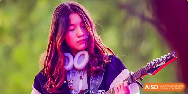 A student at Dulwich College Beijing with headphones plays guitar, outdoors with a soft-focus green background.