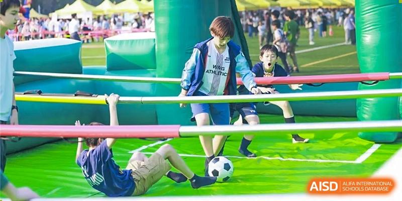 A student at Dulwich College Beijing in a blue jacket plays soccer on a field with an inflatable obstacle in the background.