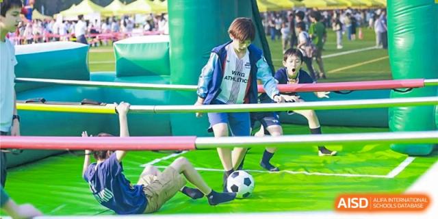 A student at Dulwich College Beijing in a blue jacket plays soccer on a field with an inflatable obstacle in the background.