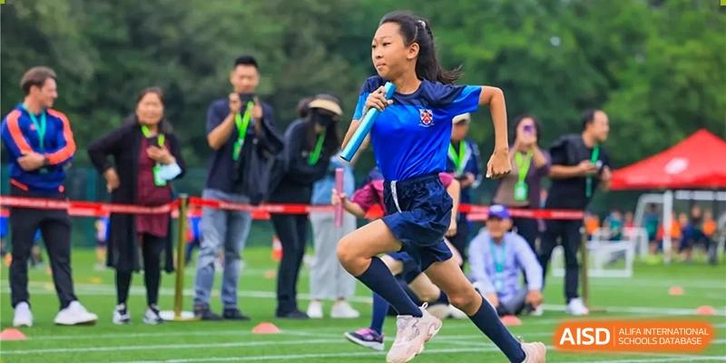 A young girl in a blue athletic uniform from Dulwich College Beijing is running a race.