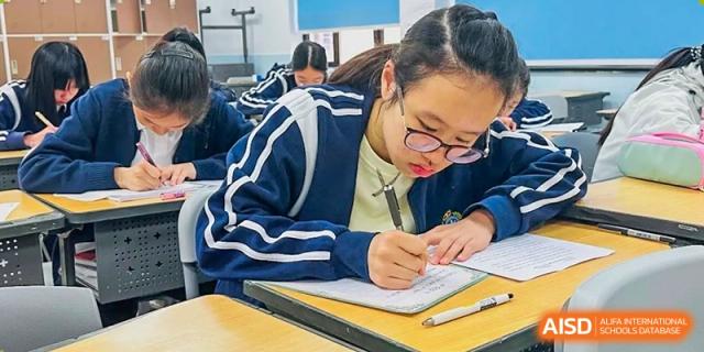 A female student in a blue jacket focused on writing notes or taking a test in a classroom at The No.2 High School Of East China Normal University (HSEFZ).