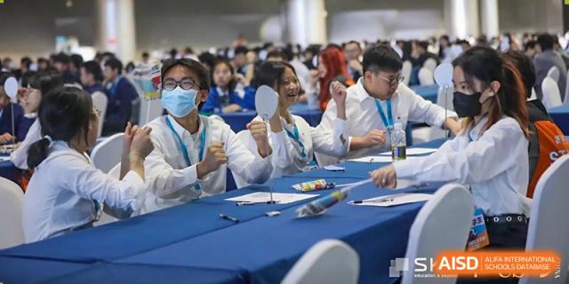 A close-up of students in white shirts and lanyards raising their thumbs up while seated at a blue-covered table at The No.2 High School Of East China Normal University (HSEFZ).