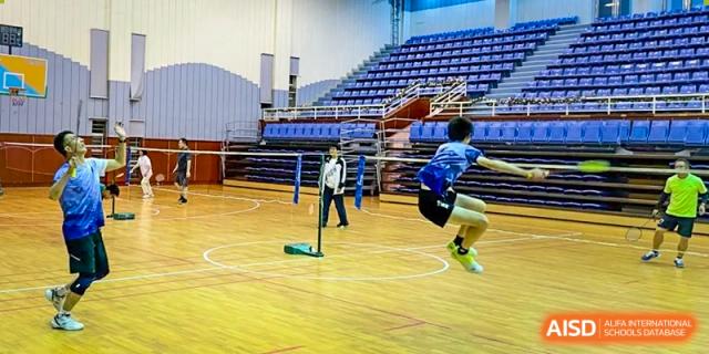 A student jumping to hit a badminton shuttlecock in the school's indoor gymnasium with large seating bleachers at The No.2 High School Of East China Normal University (HSEFZ).