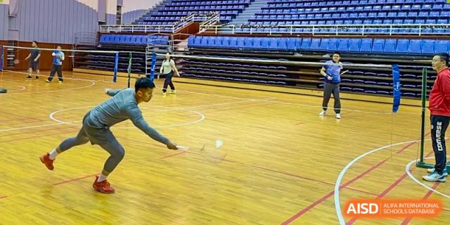 Two students playing badminton on the wooden floor of the indoor sports hall with seating bleachers in the background at The No.2 High School Of East China Normal University (HSEFZ).