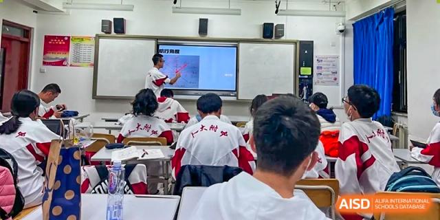 Students in red and white uniforms are seated at desks, facing the front of a classroom with a smart board at the International Curriculum Center of RDFZ.
