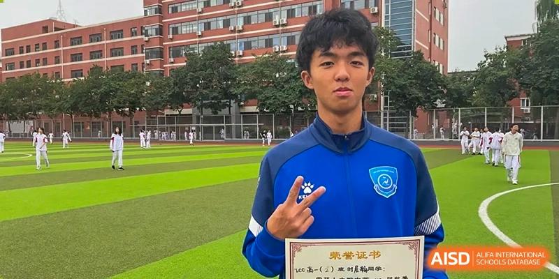 A male student in a blue and white uniform is posing for a photo, holding up a certificate at the International Curriculum Center of RDFZ.