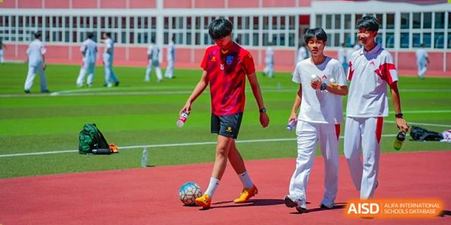 A young man in a red t-shirt and shorts is dribbling a soccer ball on a sports field at the International Curriculum Center of RDFZ.