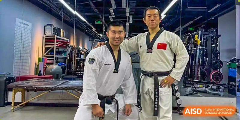 Two men, one wearing a martial arts uniform and the other a black uniform with a Chinese flag patch, are posing together in a gym at the International Curriculum Center of RDFZ.