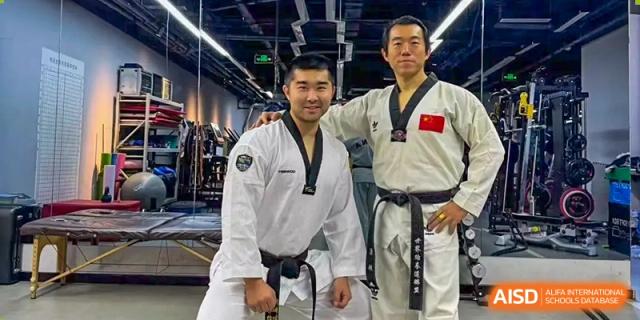 Two men, one wearing a martial arts uniform and the other a black uniform with a Chinese flag patch, are posing together in a gym at the International Curriculum Center of RDFZ.