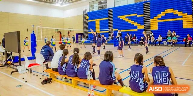A group of students from ISA Science City International School is sitting on a bench while watching a game of volleyball.