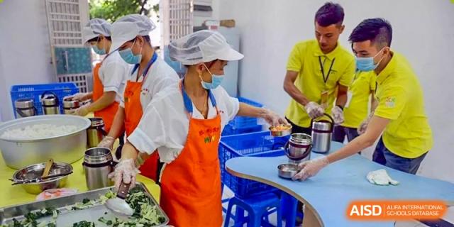 A kitchen staff member from ISA Science City International School, wearing an orange apron and a hairnet, is preparing food.