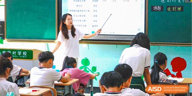 A female teacher in a white dress teaching young students who are sitting on the floor in front of a whiteboard at Maple Leaf Foreign Nationals School-Yiwu.