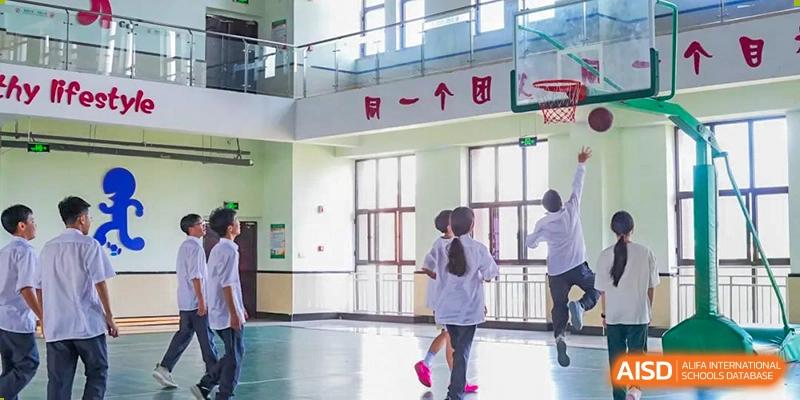 Students playing basketball indoors near a hoop and a second-floor glass railing at Maple Leaf Foreign Nationals School-Yiwu.