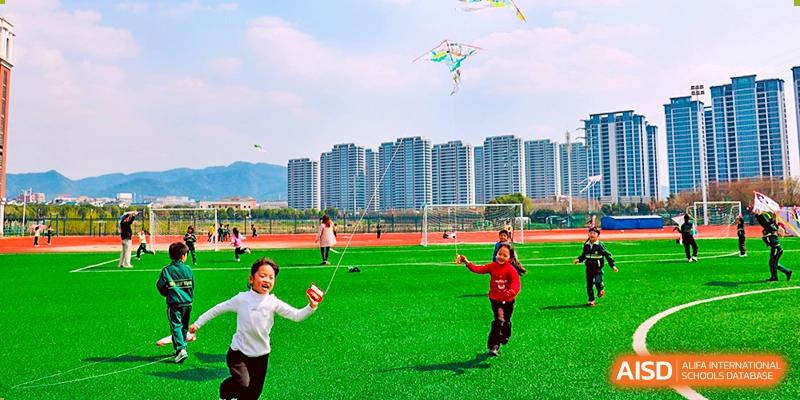 Happy students flying a kite and playing on a bright green field with a backdrop of tall apartment buildings near Maple Leaf Foreign Nationals School-Yiwu.