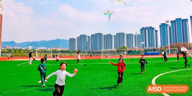 Happy students flying a kite and playing on a bright green field with a backdrop of tall apartment buildings near Maple Leaf Foreign Nationals School-Yiwu.