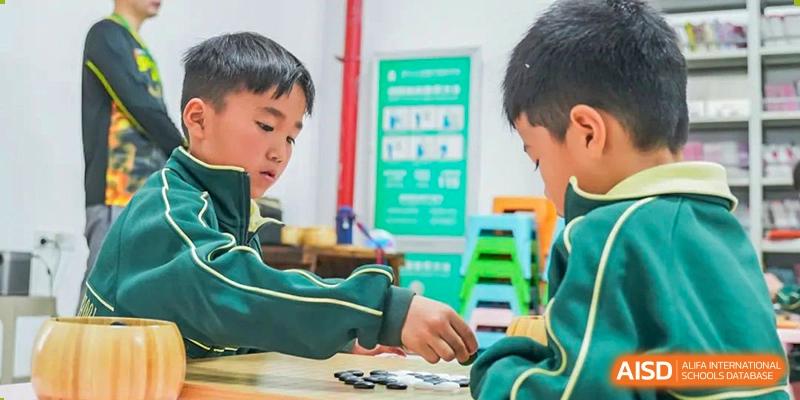 A male student in a green uniform works on a project or task at a desk in a classroom setting at Maple Leaf Foreign Nationals School-Yiwu.
