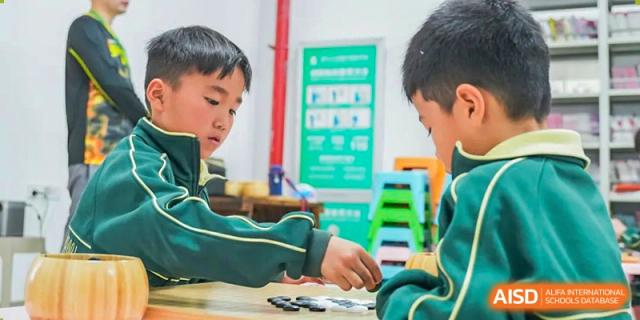 A male student in a green uniform works on a project or task at a desk in a classroom setting at Maple Leaf Foreign Nationals School-Yiwu.