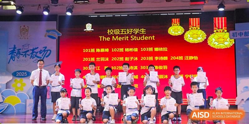 Students standing on a brightly lit stage holding certificates under a banner that reads "The Merit Student" at Maple Leaf Foreign Nationals School-Yiwu.