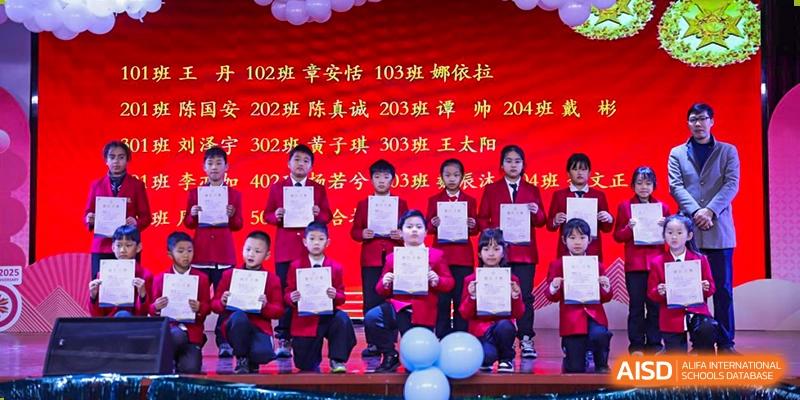 A group of young students in red shirts holding certificates and standing with blue balloons on a stage against a red background with white and yellow text at Maple Leaf Foreign Nationals School-Yiwu.