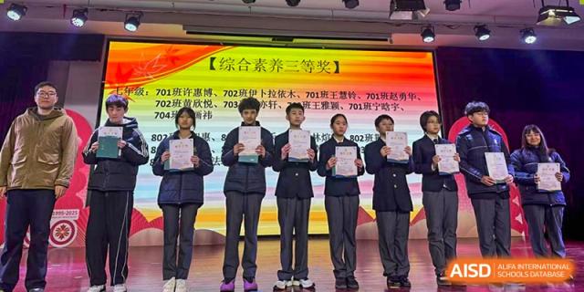 Students in dark uniforms holding up certificates of achievement on a stage with a banner displaying Chinese text and a "Second Prize" designation at Maple Leaf Foreign Nationals School-Yiwu.