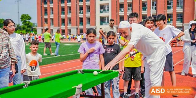Students and an older man playing a game of pool/billiards on an outdoor red track near a school building at Maple Leaf Foreign Nationals School-Yiwu.