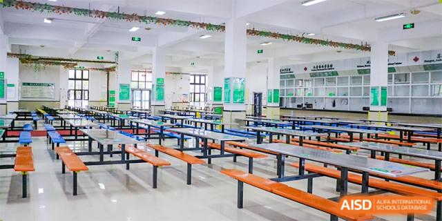 A large, empty school cafeteria with rows of tables and attached orange benches, ready for meal service at Maple Leaf Foreign Nationals School-Yiwu.