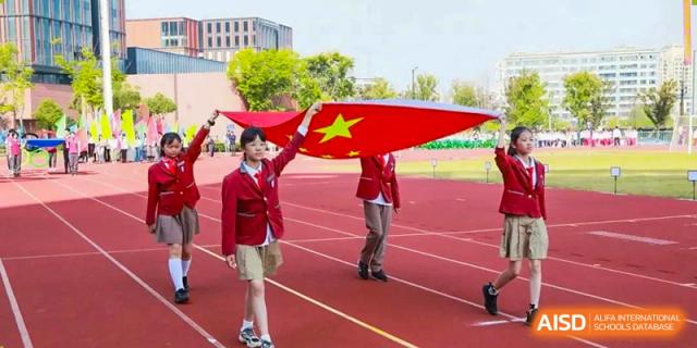 Two NANJING THOMAS SCHOOL students are holding and waving a large Chinese flag on a sports field.