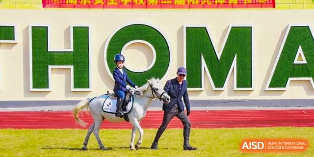 A NANJING THOMAS SCHOOL student is riding a horse, with an adult dressed as a jockey walking next to them.