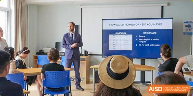A teacher in a suit presenting to a student wearing a straw hat in a classroom at the Qingdao Amerasia International School.