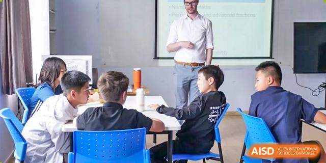 A teacher standing in front of a whiteboard and a young male student in a blue chair, who is raising his hand at the Qingdao Amerasia International School.