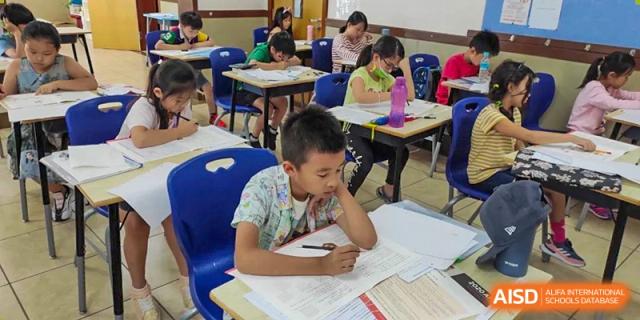 A young male student with a pensive expression writing on a paper at his desk in a classroom at the Qingdao Amerasia International School.