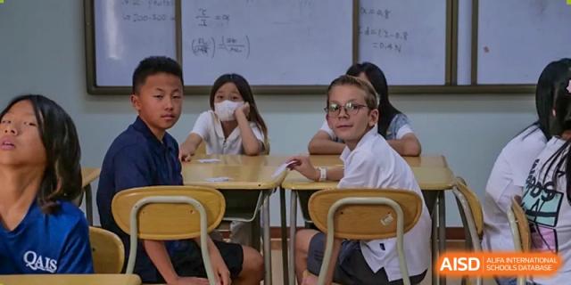 Two students with glasses sitting at desks in a classroom, looking towards the front of the room at the Qingdao Amerasia International School.