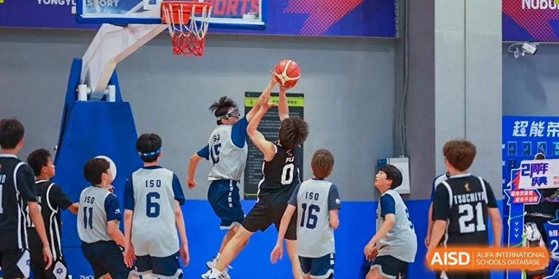 Two teenage boys jumping to block a shot during a basketball game, with one player attempting to lay up the ball at the Qingdao Amerasia International School.