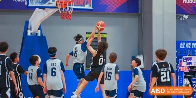 Two teenage boys jumping to block a shot during a basketball game, with one player attempting to lay up the ball at the Qingdao Amerasia International School.